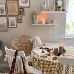 Klaudia Makowska-Kuźma - Stół nakryty żółto-białym obrusem w kratę, dekorowany liśćmi i świecami, z otwartą książką, w tle rattanowy fotel i półki z dekoracjami.