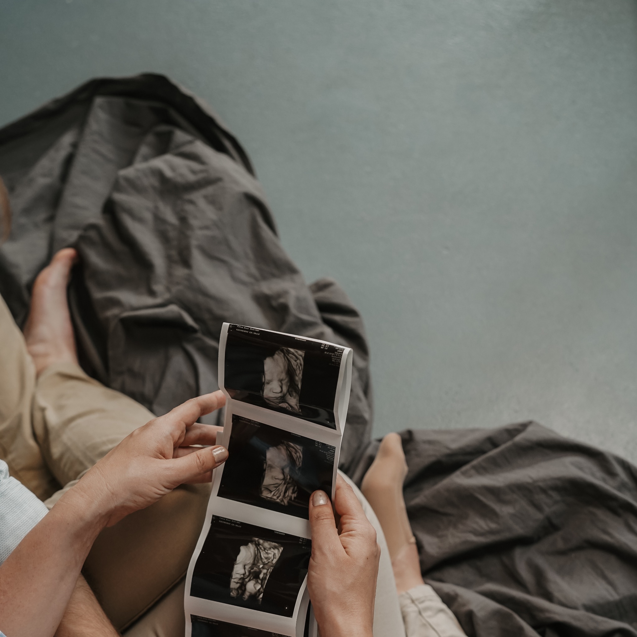 Rodzice siedzą na łóżku, trzymając w rękach serię wydruków USG dziecka, z widocznymi szczegółami twarzy nienarodzonego dziecka.