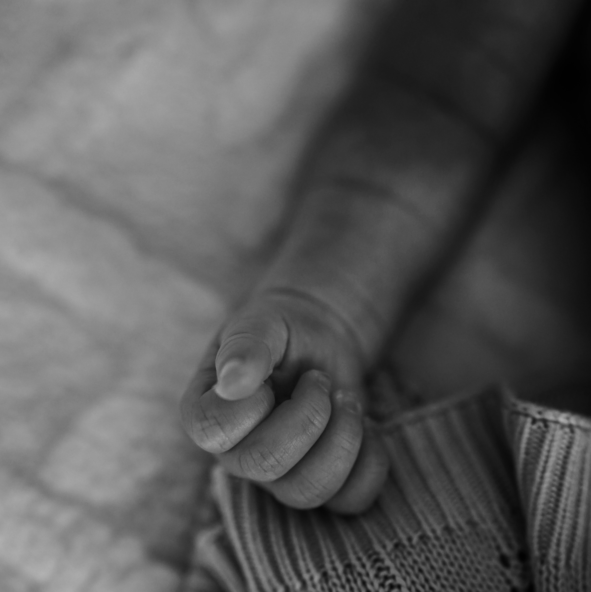 Dłoń noworodka, uchwycona w czerni i bieli, z delikatnymi fałdkami skóry i drobnymi palcami, spoczywająca na miękkiej, dzierganej tkaninie w paski.