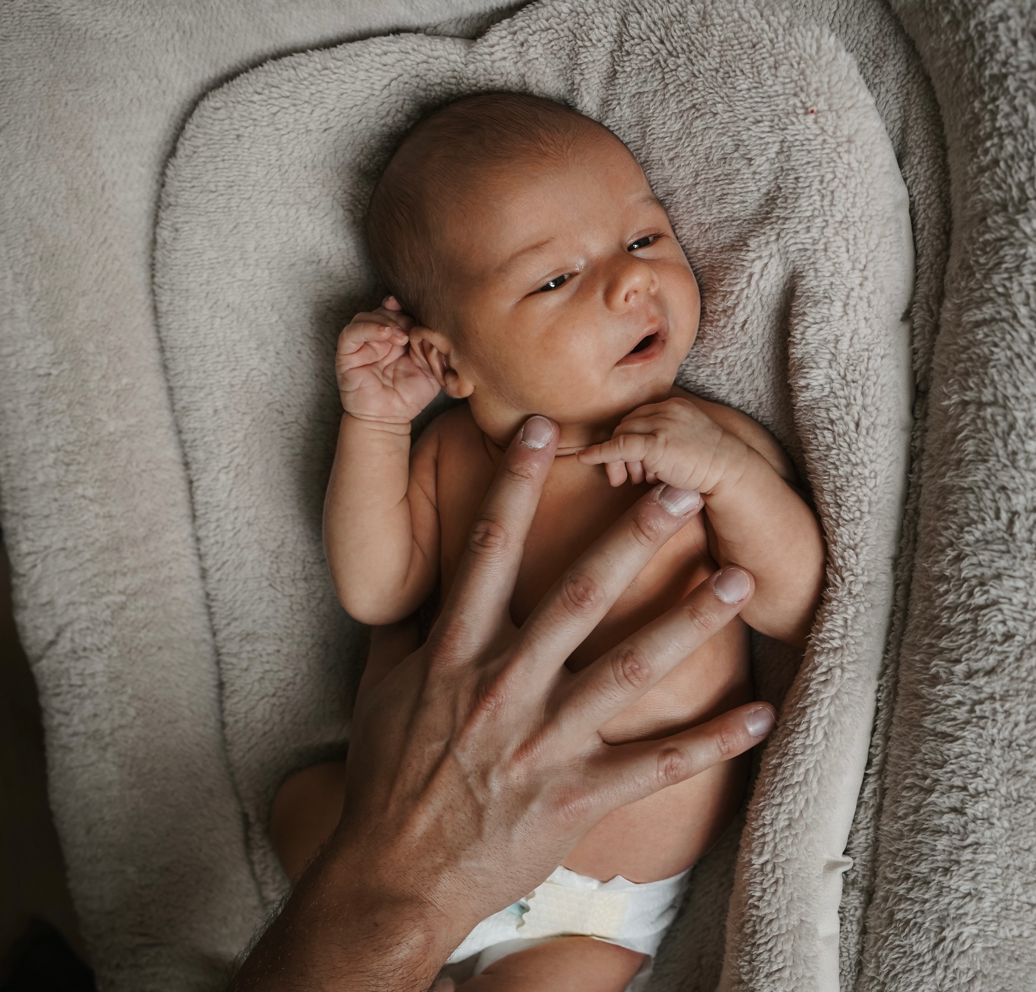 Noworodek leży na miękkim, beżowym kocyku, delikatnie dotykany przez dłoń dorosłego. Dziecko ma otwarte oczy i wyraz zaciekawienia na twarzy.