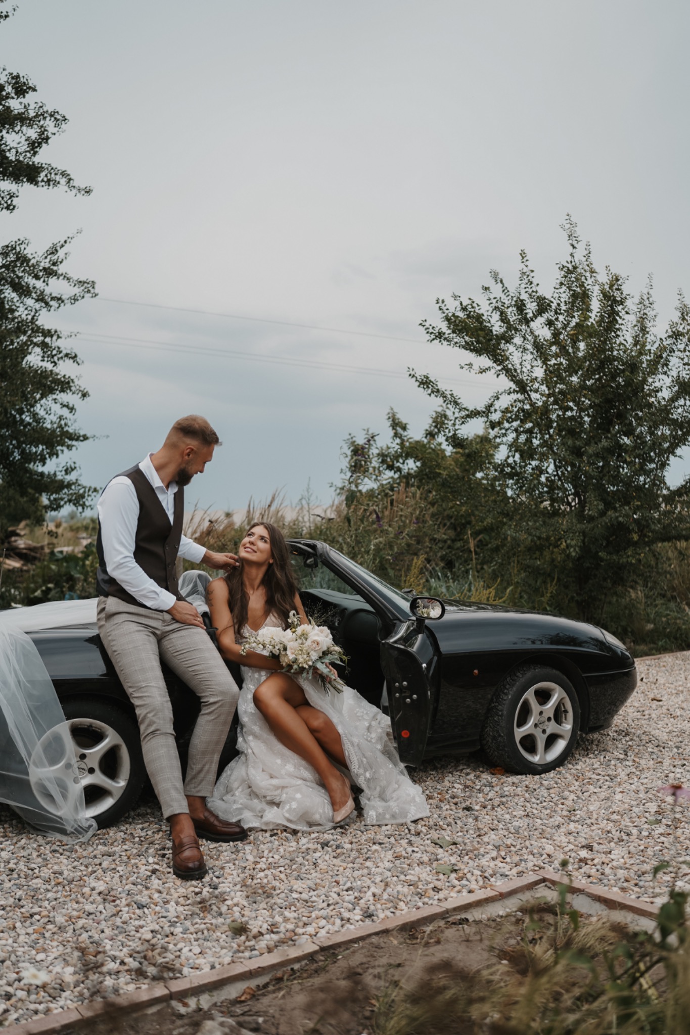 Para młoda pozuje przy czarnym kabriolecie na tle zieleni, panna młoda trzyma bukiet białych róż, a pan młody opiera się o samochód.