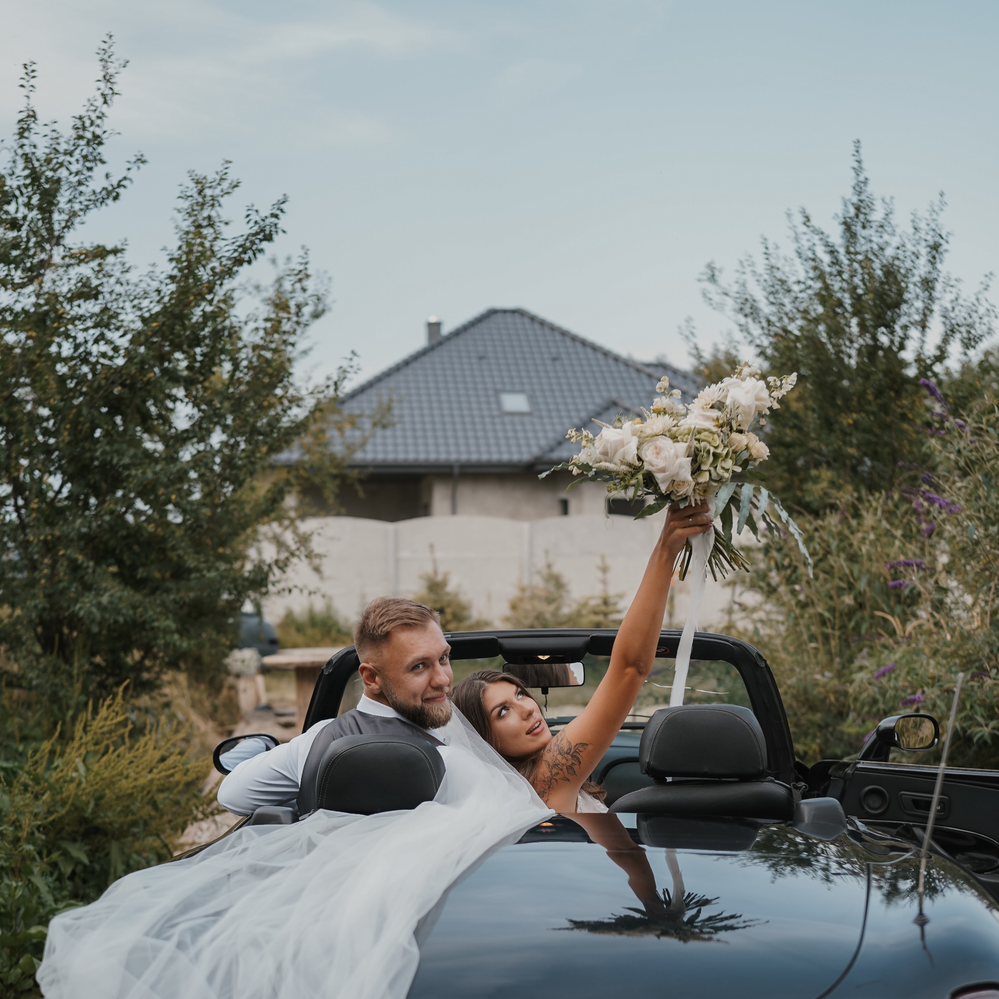 Para młoda w kabriolecie: panna młoda trzyma bukiet białych róż, widoczny tatuaż na ramieniu, w tle dom i zieleń.