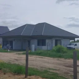 Niedokończony dom jednorodzinny z ciemną dach&oacute;wką, widoczny zza drewnianego ogrodzenia, z zaparkowanym białym vanem w tle.