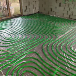 Instalacja ogrzewania podłogowego z zielonych rurek na srebrnej folii, widok z góry na surowy stan budynku z oknem w tle.