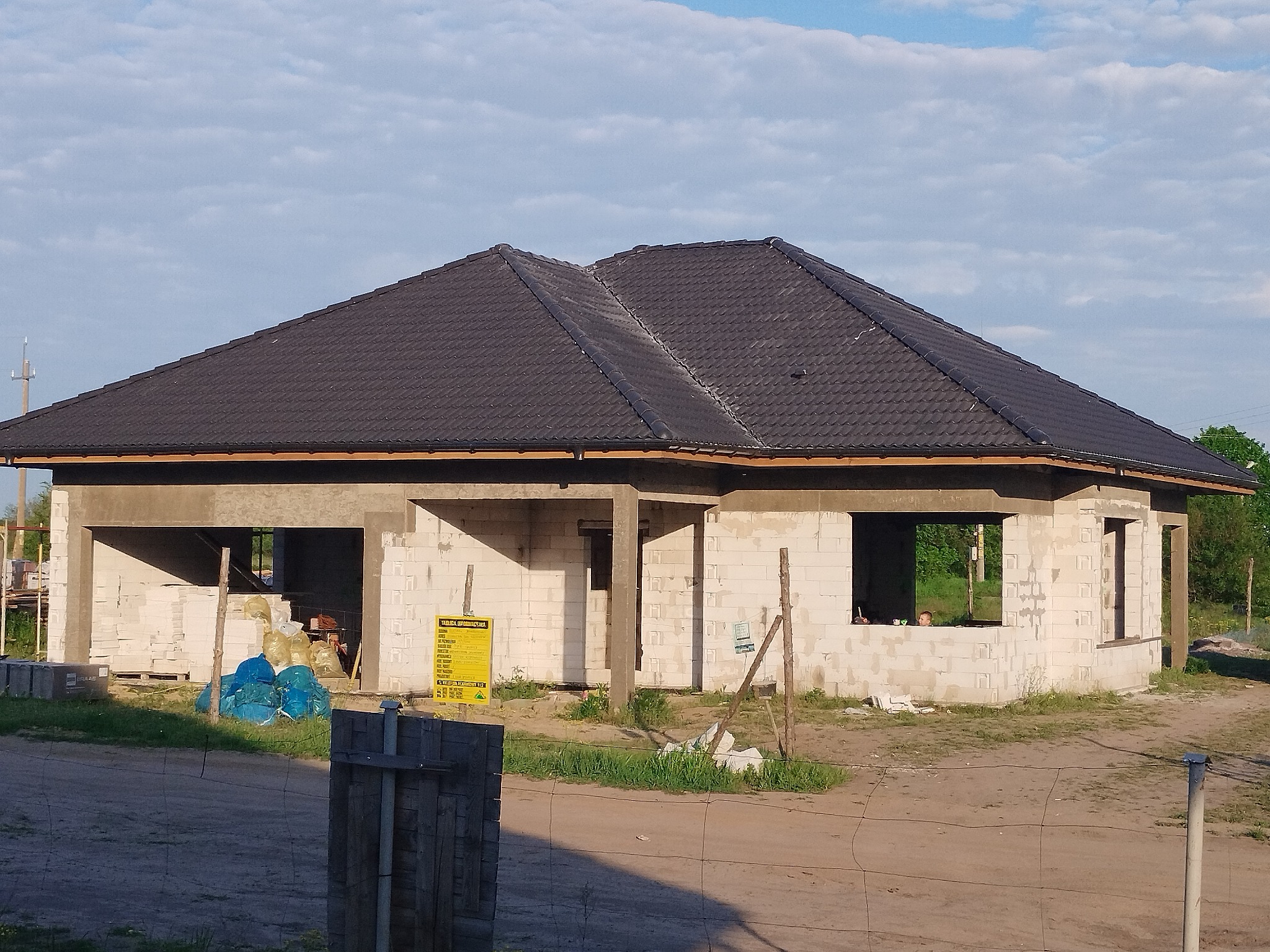 Surowy stan domu jednorodzinnego w trakcie budowy, widoczne ściany z bloczków, czarny dach i materiały budowlane na posesji.
