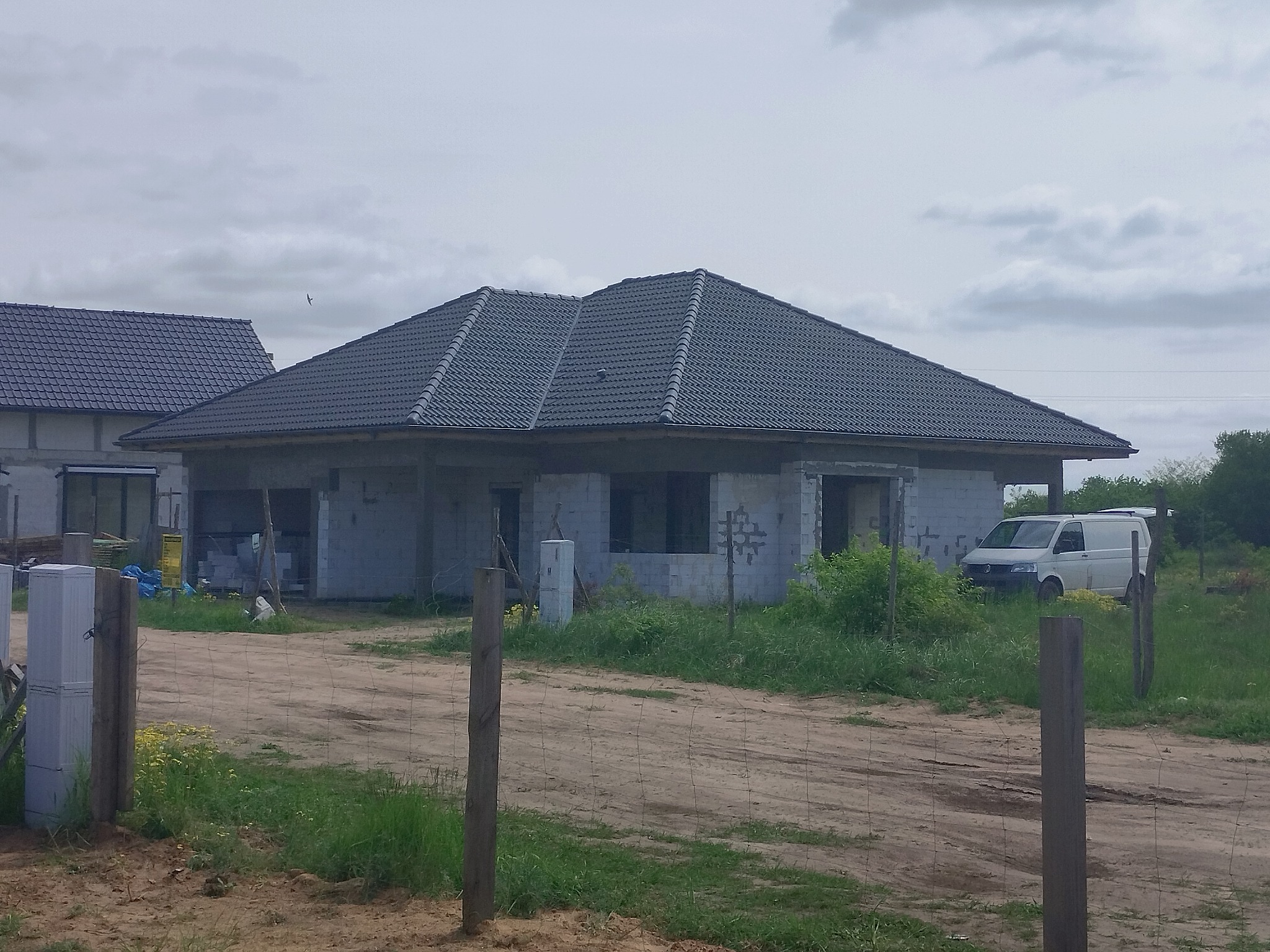 Niedokończony dom jednorodzinny z ciemną dachówką, widoczny zza drewnianego ogrodzenia, z zaparkowanym białym vanem w tle.