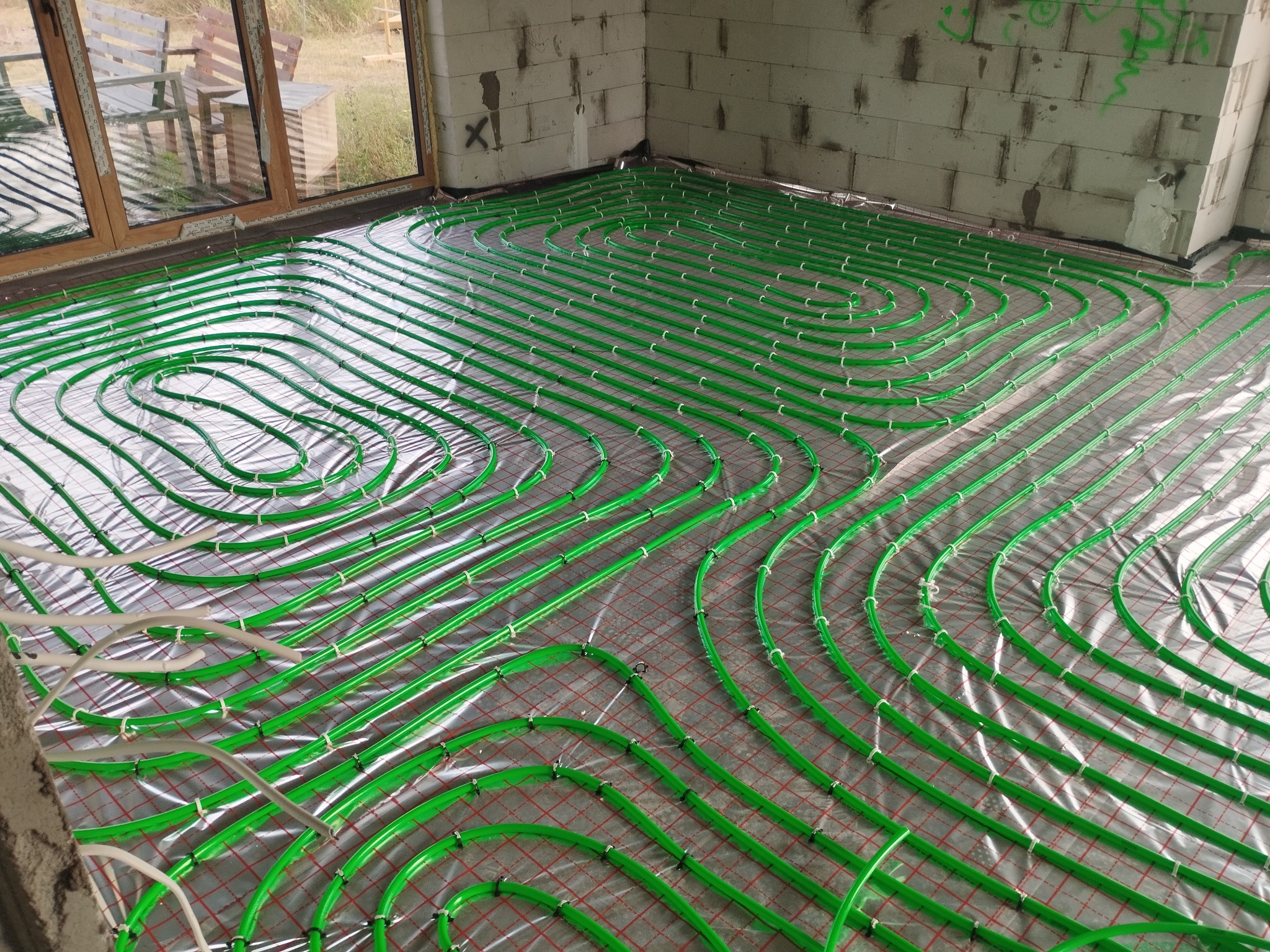 Instalacja ogrzewania podłogowego z zielonych rurek na srebrnej folii, widok z góry na surowy stan budynku z oknem w tle.