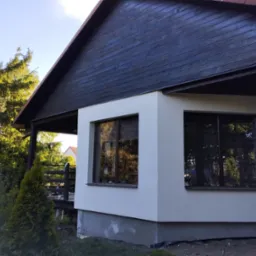 Fragment domu z oknami po wykonaniu elewacji, widoczna gładka, jasna powierzchnia ściany i ciemne drewniane wykończenie górnej części budynku.
