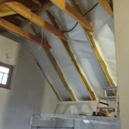 Poddasze w trakcie ocieplania wełną mineralną, widoczne drewniane krokwie, biała membrana paroizolacyjna, rusztowanie i drabina oparta o ścianę, okno w szarej ścianie.
