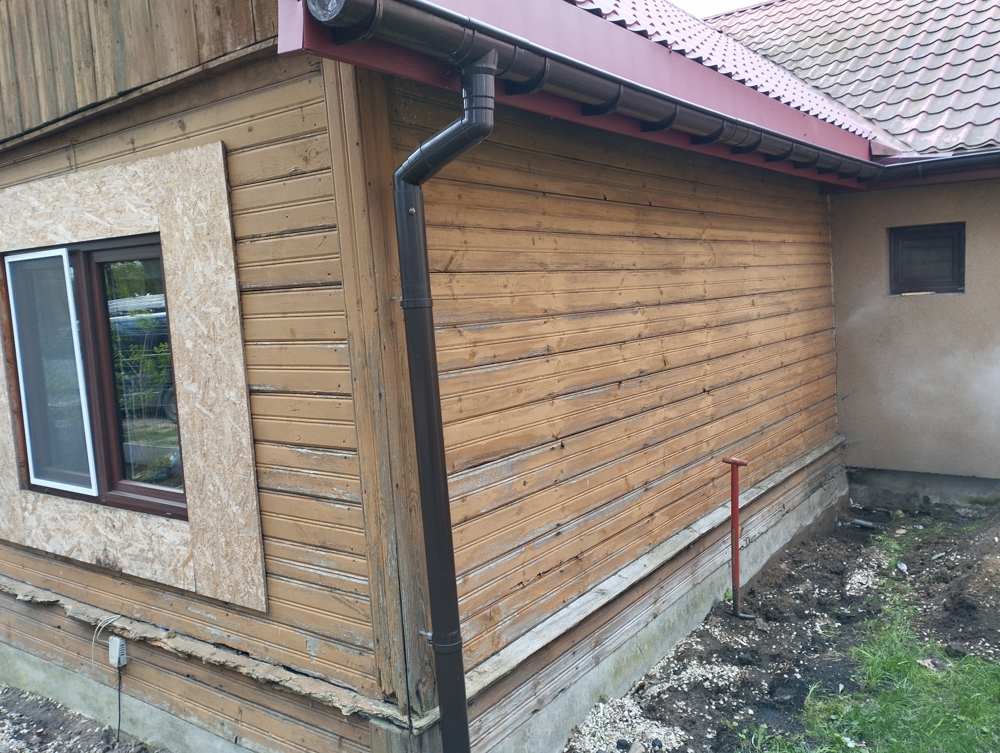 Fragment drewnianego domu z brązową rynną, częściowo obłożonego płytą OSB w miejscu okna, z widocznymi uszkodzeniami elewacji i czerwoną łopatą w ziemi.