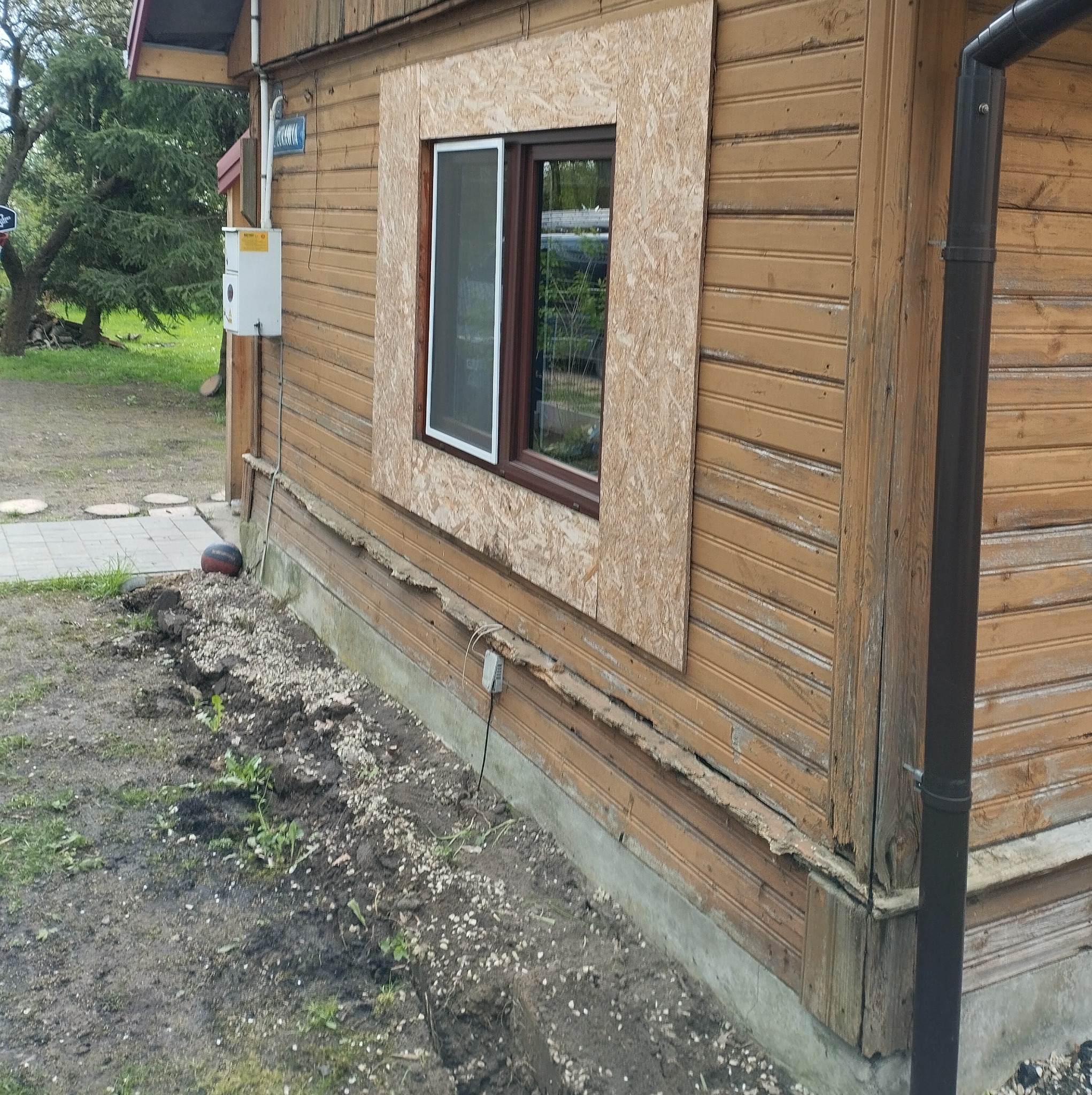 Drewniany dom z brązowymi deskami, okno zabezpieczone płytą OSB, widoczne ślady uszkodzeń elewacji i przygotowanie do prac ziemnych wzdłuż fundamentu.