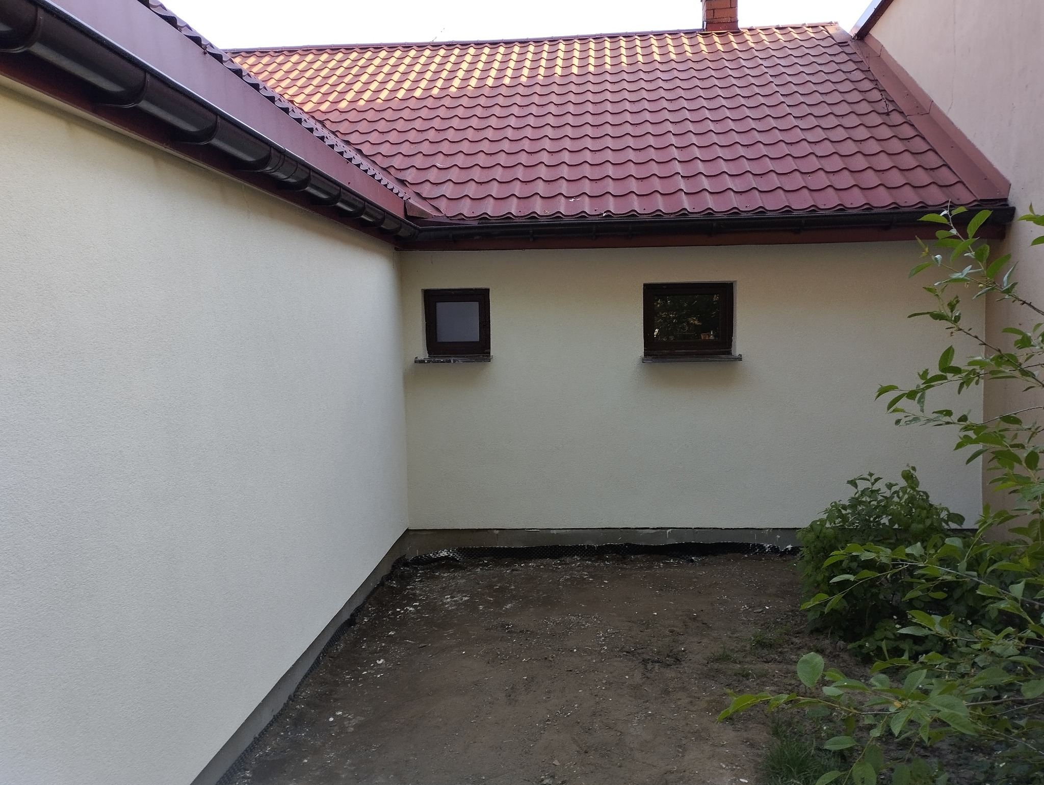 Świeżo otynkowana ściana zewnętrzna budynku z dwoma małymi oknami i nowym orynnowaniem, widoczne elementy drenażu przy gruncie, czerwona dachówka.