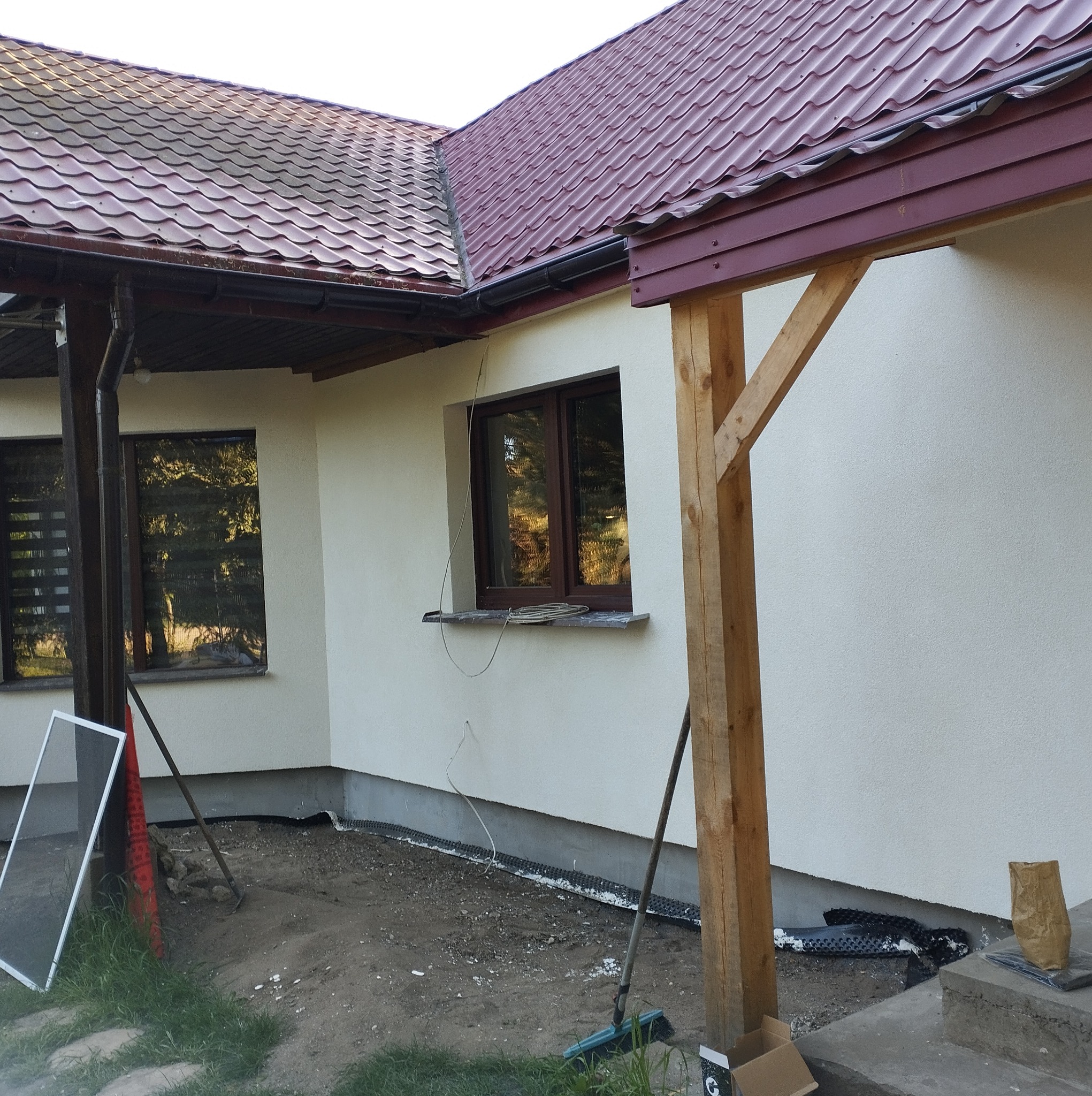 Dom jednorodzinny z jasną elewacją, brązowymi oknami i ciemnoczerwoną dachówką, widoczne przygotowania do prac wykończeniowych wokół budynku, w tym rozłożona folia fundamentowa i narzędzia.