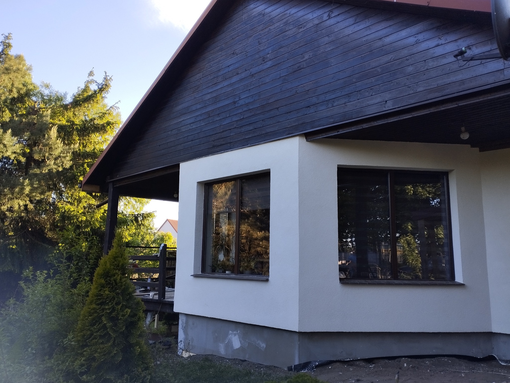 Fragment domu z oknami po wykonaniu elewacji, widoczna gładka, jasna powierzchnia ściany i ciemne drewniane wykończenie górnej części budynku.