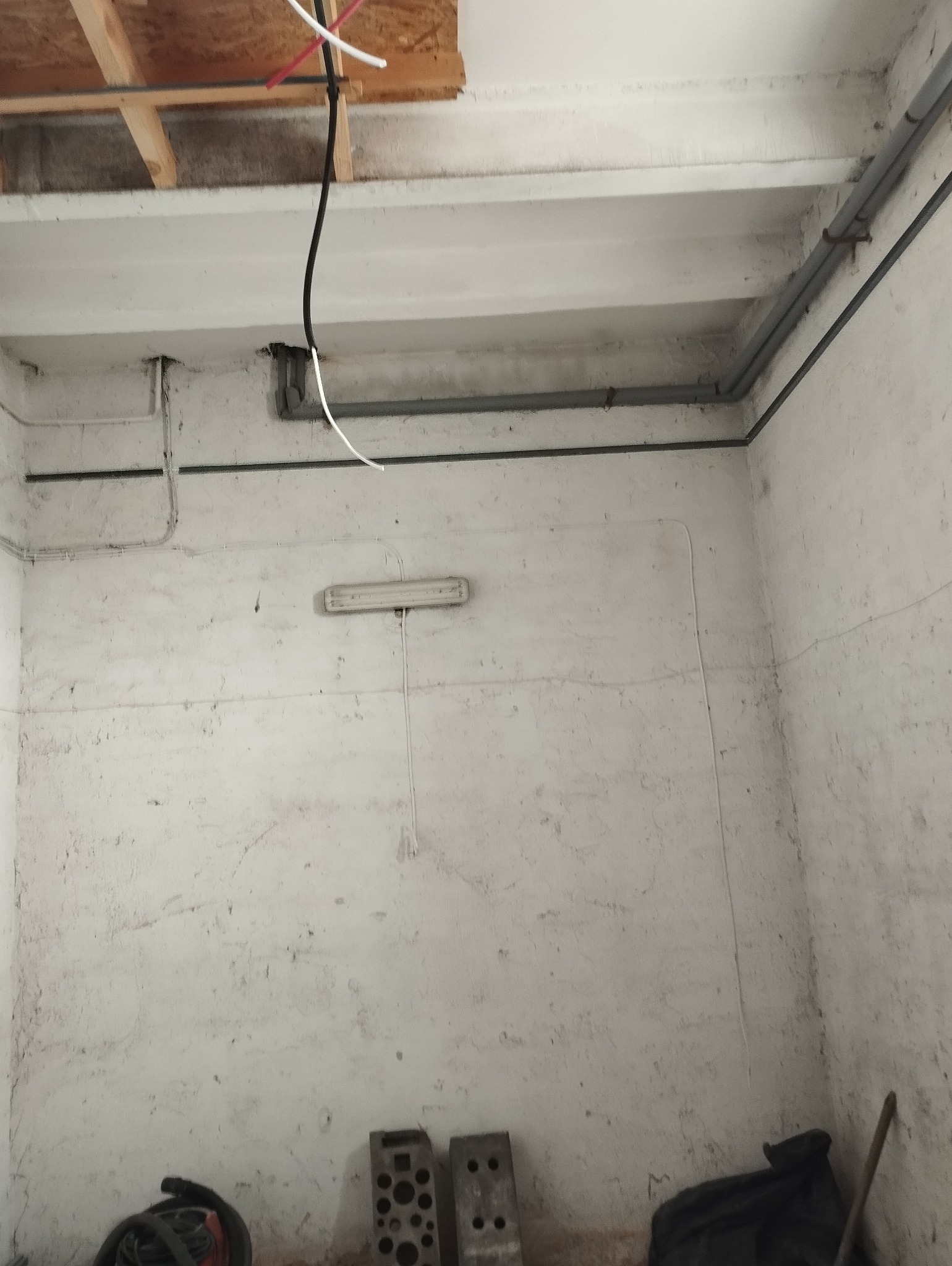 Surowe wnętrze z widocznymi przewodami elektrycznymi, rurami i lampą fluorescencyjną na nierównej, pomalowanej na biało ścianie. Na podłodze widoczne narzędzia i gruz.