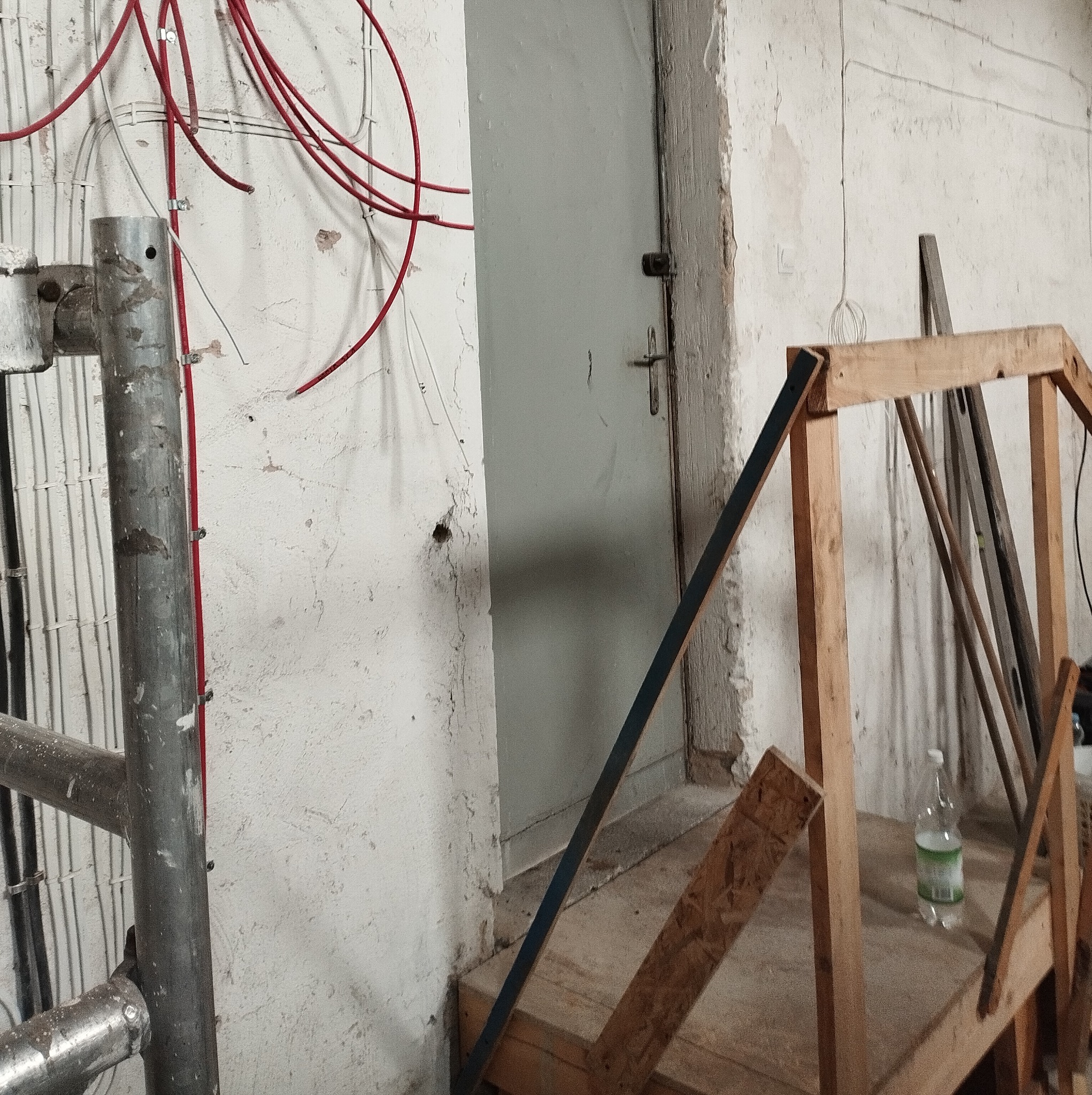 Wnętrze w trakcie prac remontowych: widoczne przewody elektryczne na ścianie, metalowe rusztowanie, drewniana konstrukcja, butelka wody i stara, szara, metalowa futryna drzwiowa.