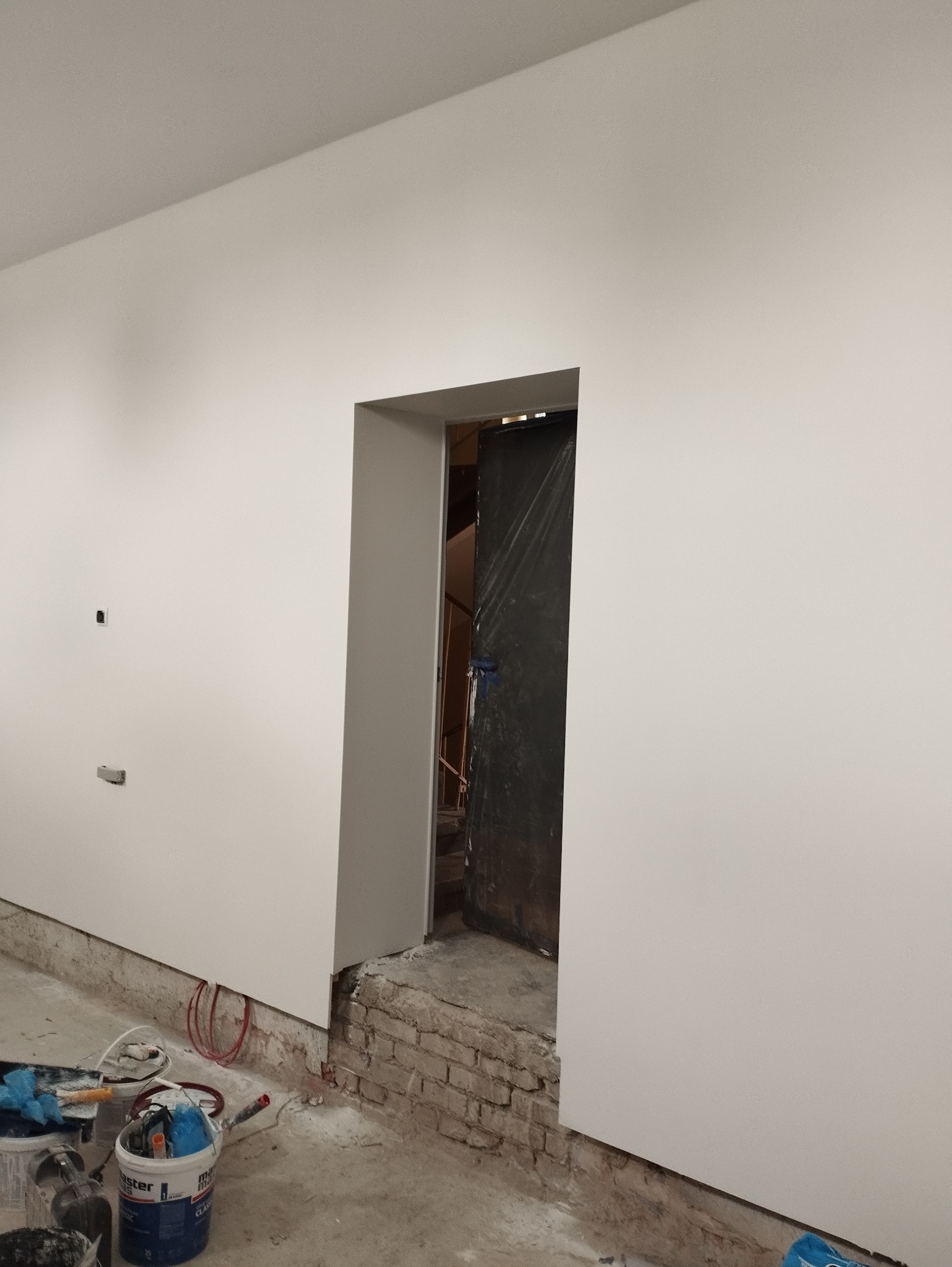 Świeżo pomalowana na biało ściana z widocznymi puszkami z farbą i narzędziami malarskimi na podłodze, zarys ceglanej podstawy ściany oraz zasłonięte folią drzwi w tle.