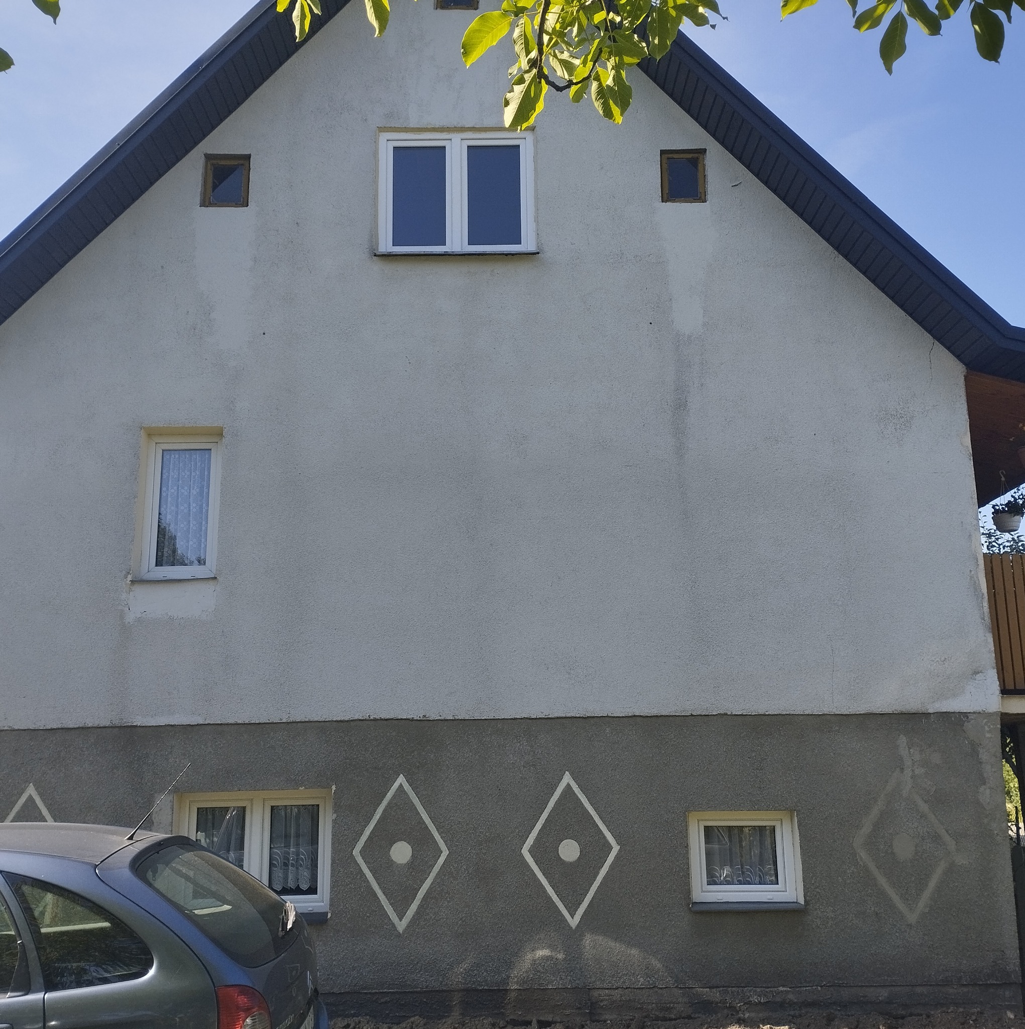 Dwukondygnacyjny dom z szarą elewacją, białymi ramami okiennymi i ozdobnymi rombami w dolnej części; zaparkowany samochód w lewym dolnym rogu.