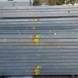 Józef Bobula - P.P.H.U BUD-MAX - Stos szarych, metalowych profili o prostokątnym przekroju, ułożonych na zewnątrz, widoczne żółte etykiety, w tle resztki śniegu i budynek.