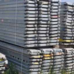 Józef Bobula - P.P.H.U BUD-MAX - Stosy srebrnych, metalowych platform roboczych ułożonych na paletach, widoczne ślady użytkowania i niewielkie plamy farby, na zewnątrz, w tle zieleń.