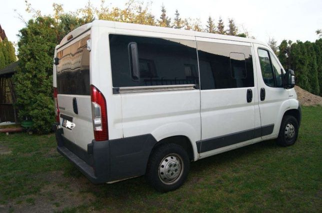Biały bus dostawczy z przesuwnymi drzwiami, zaparkowany na trawie przed domem z ogrodem.