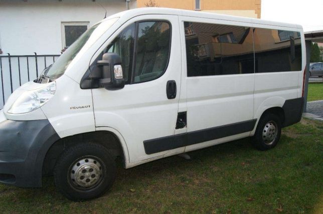 Biały bus marki Peugeot z przyciemnianymi szybami, zaparkowany na trawniku przed budynkiem.
