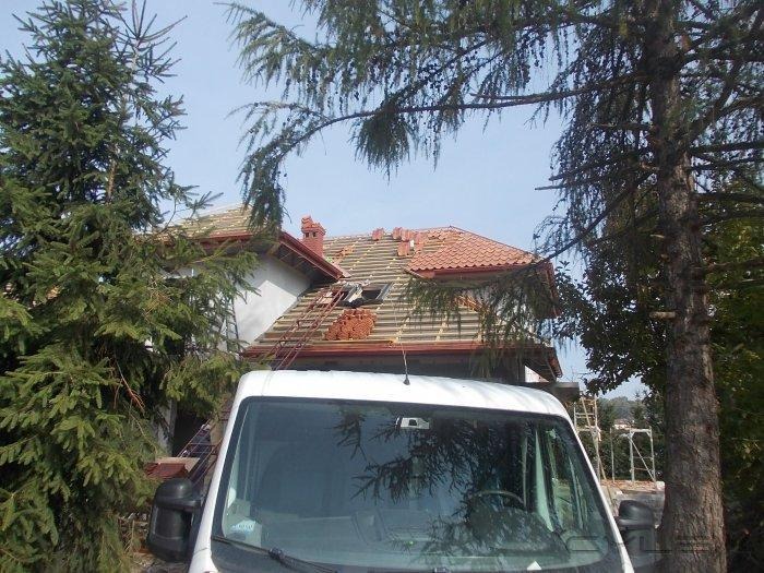 Dom w trakcie remontu dachu, widoczna część starej dachówki, nowa dachówka na palecie na dachu i drabina oparta o dach, biały bus na pierwszym planie.