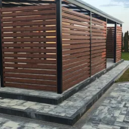 Nowoczesna, brązowa przesłona z poziomych listew zamontowana na czarnej, metalowej konstrukcji z przezroczystym zadaszeniem, otaczająca taras domu z żółtą elewacją, widoczna z poziomu kostki brukowej.