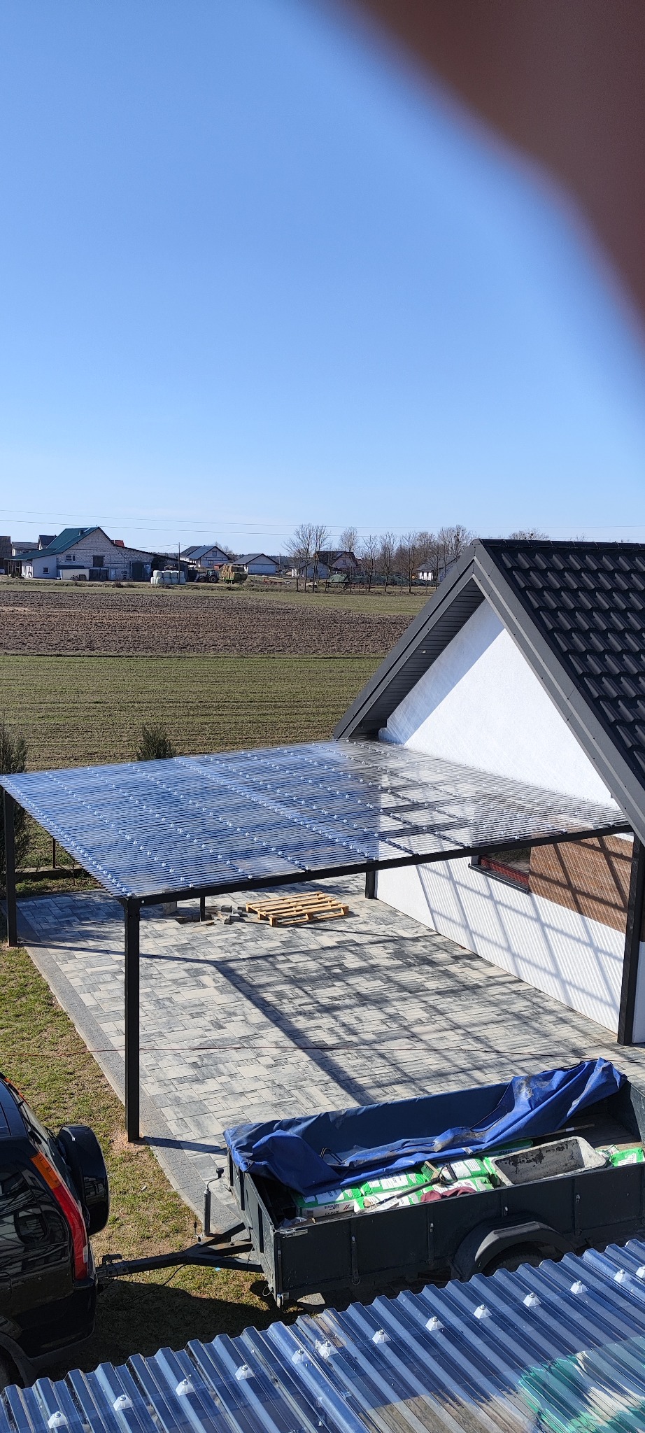 Zadaszenie tarasu z przezroczystego poliwęglanu na metalowej konstrukcji, widok z góry na posesję z kostką brukową, przyczepką i samochodem w tle pola uprawne i zabudowania.
