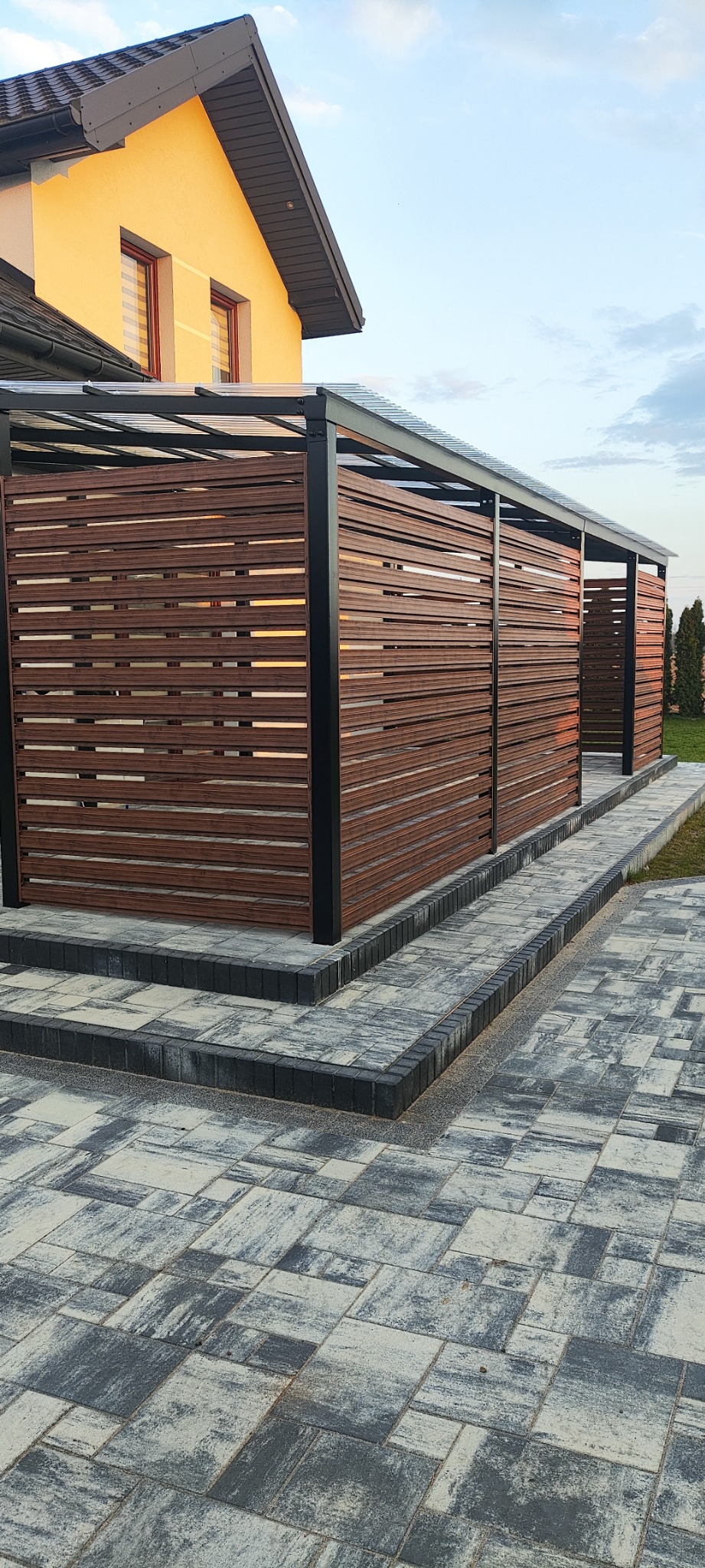 Nowoczesna, brązowa przesłona z poziomych listew zamontowana na czarnej, metalowej konstrukcji z przezroczystym zadaszeniem, otaczająca taras domu z żółtą elewacją, widoczna z poziomu kostki brukowej.