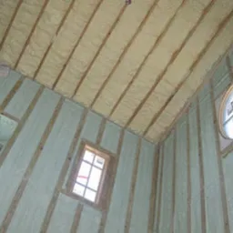 Poddasze z drewnianą konstrukcją szkieletową wypełnione jasną pianką izolacyjną, widoczne okno okrągłe i prostokątne.