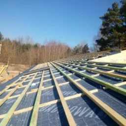 Konstrukcja dachu w trakcie budowy: widoczne membrany dachowe z nadrukiem i drewniane łaty pod dachówki, na tle błękitnego nieba i drzew.