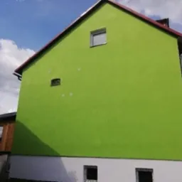 Ściana szczytowa domu jednorodzinnego z jaskrawozieloną elewacją, widoczne ślady po mocowaniu rusztowania, biały cokół i czerwona dachówka.