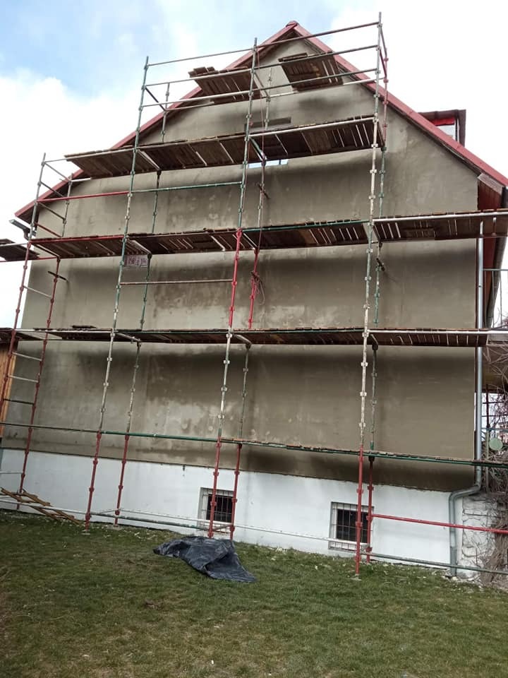 Dom w trakcie prac elewacyjnych: szare ściany z fragmentami białego podkładu, metalowe rusztowanie z drewnianymi pomostami na tle zielonego trawnika i pochmurnego nieba.