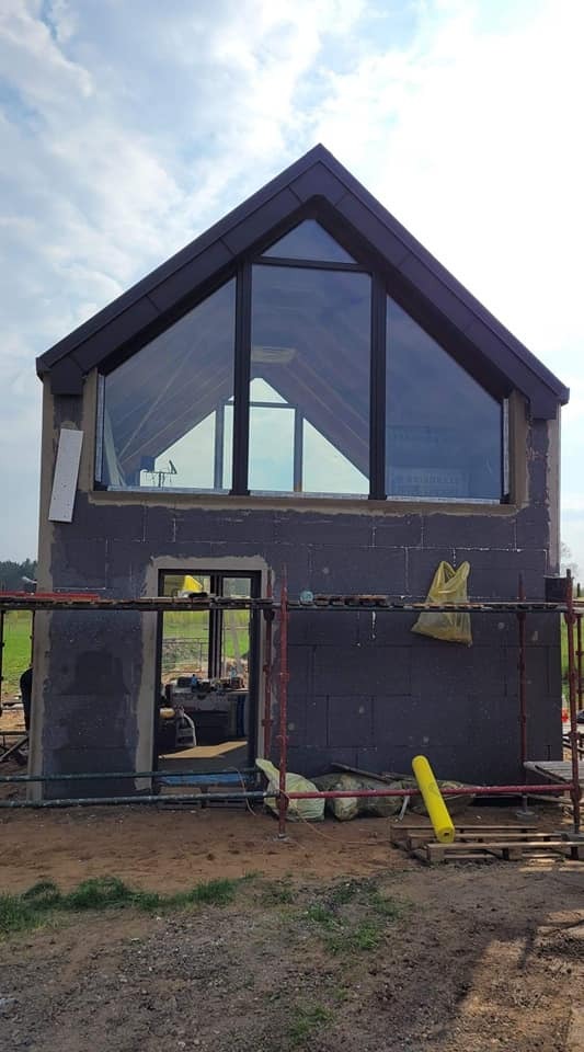 Surowy stan domu z dużym oknem trójkątnym na poddaszu, widoczne rusztowania i ocieplenie ścian zewnętrznych styropianem grafitowym w trakcie montażu, teren budowy z ziemią i narzędziami.