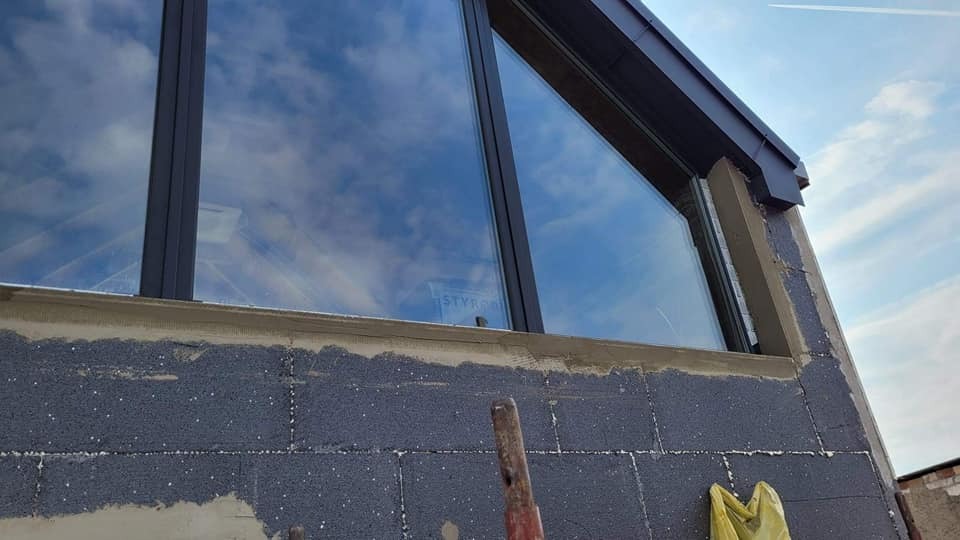 Ocieplanie elewacji budynku z oknami o nietypowym kształcie, widoczne prace przygotowawcze, częściowo położony styropian i zaprawa, tło: błękitne niebo z smugami kondensacyjnymi.