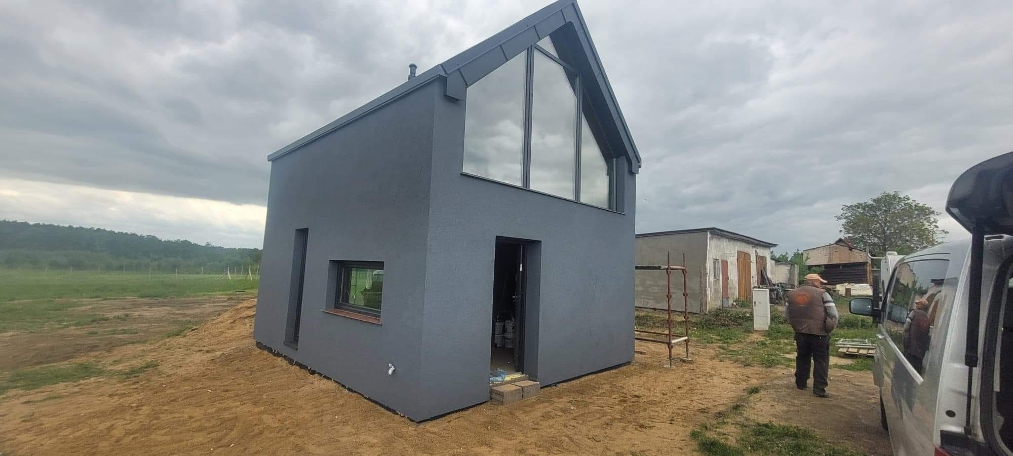 Nowoczesny, szary dom z dużym trójkątnym oknem, świeżo po nałożeniu tynku elewacyjnego, widoczny pracownik w oddali i samochód dostawczy.