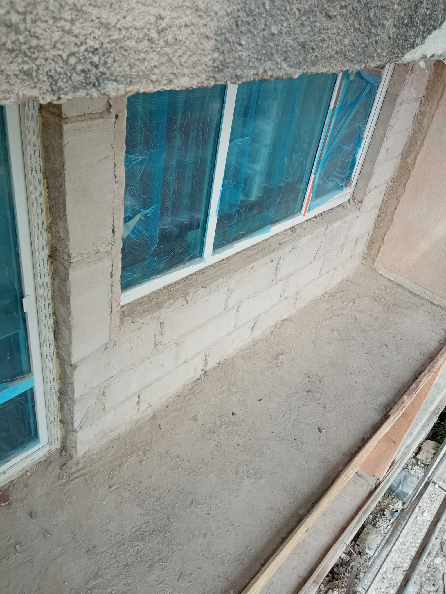Wnęka okienna w trakcie murowania z widocznymi bloczkami, zabezpieczonymi folią oknami i surowym betonowym podłożem.