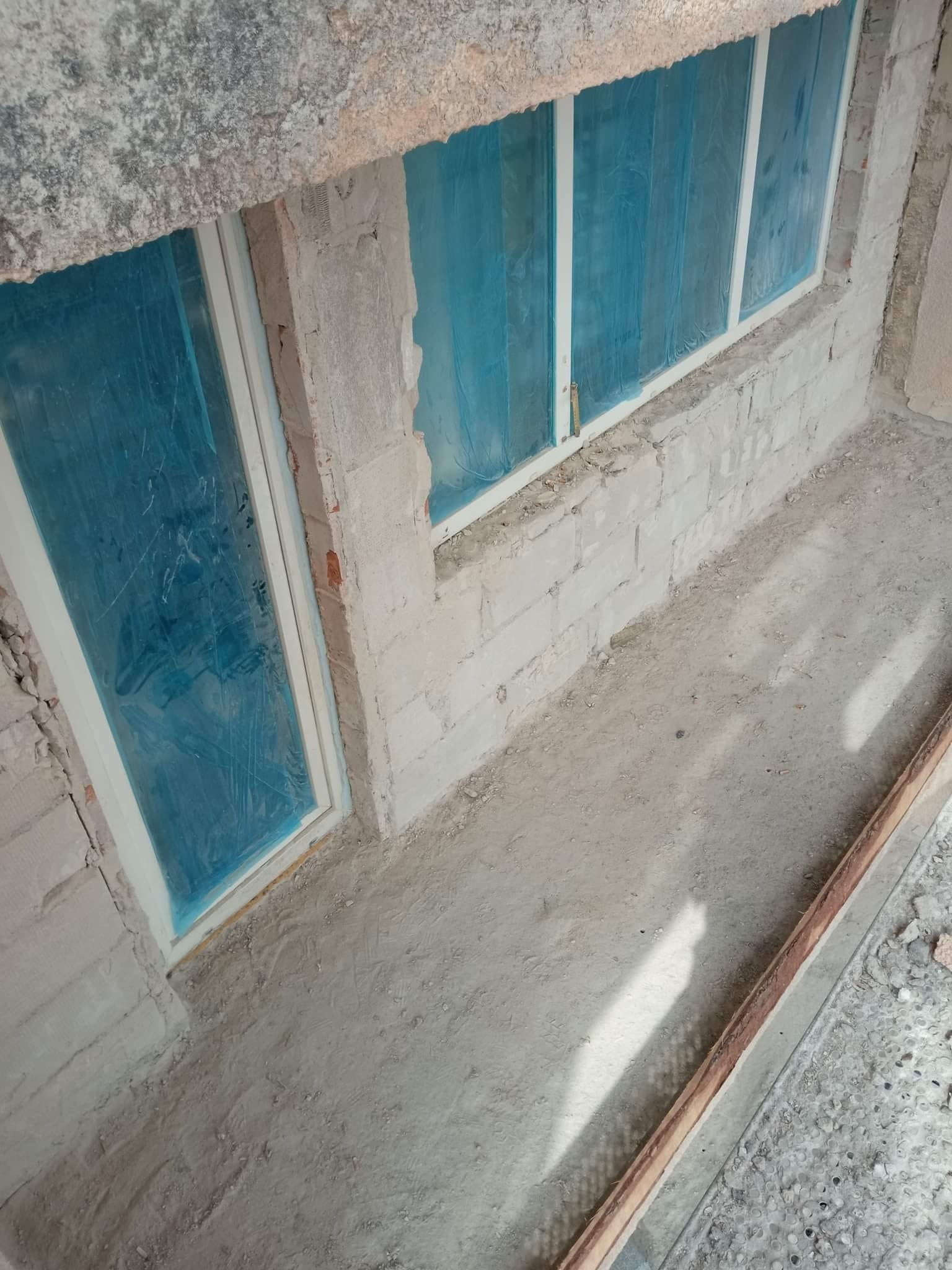 Nowe okna PCV z białymi ramami, zabezpieczone niebieską folią ochronną, osadzone w surowej ścianie z białego pustaka, przed tynkowaniem.