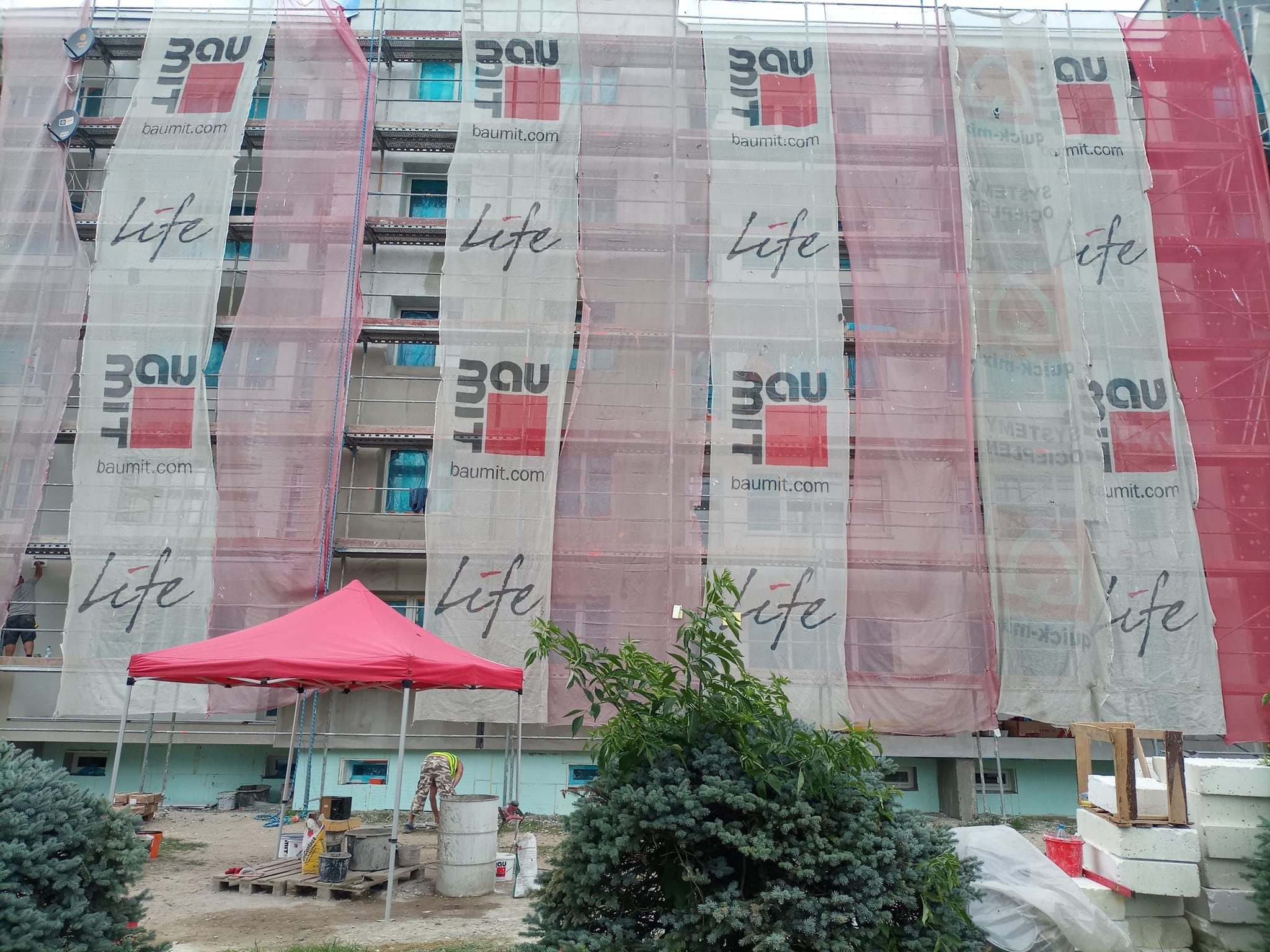 Budynek wielorodzinny w trakcie prac, osłonięty siatkami z logo Baumit Life, widoczne rusztowania i pracownicy na zewnątrz, częściowo ocieplona elewacja, na dole rozstawiony czerwony namiot.