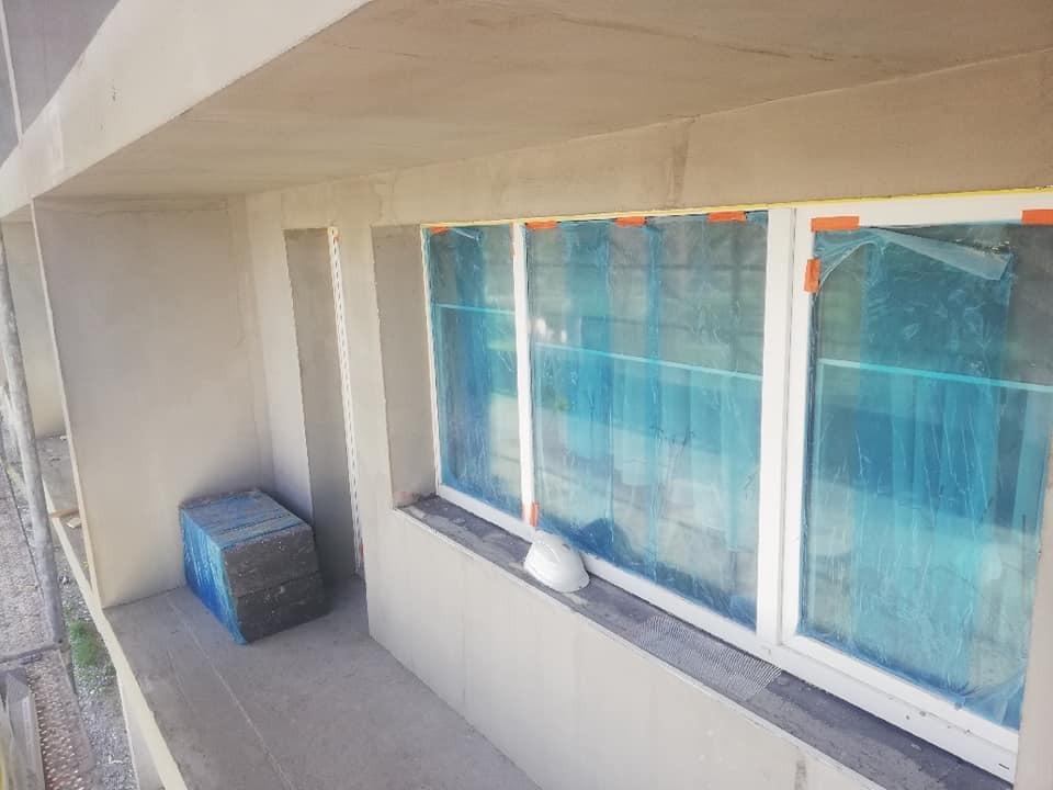 Elewacja budynku w trakcie prac wykończeniowych: okna zabezpieczone folią, ściany pokryte szarym tynkiem, na parapecie leży biały kask ochronny, a na balkonie spakowane bloczki.