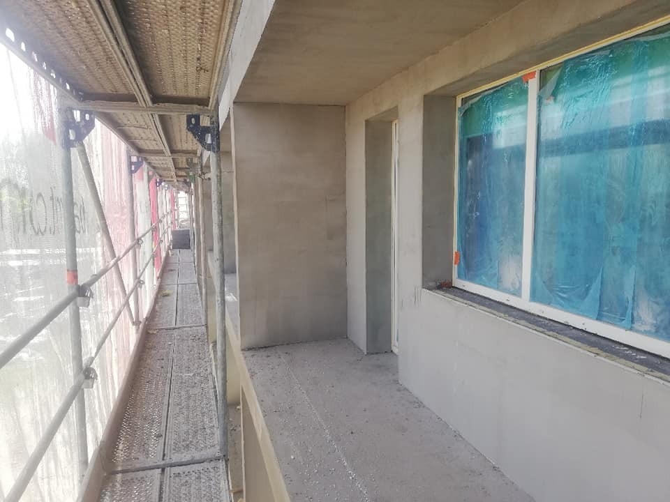 Wnętrze balkonu w trakcie prac wykończeniowych, ściany pokryte świeżym tynkiem, okno zabezpieczone folią, widoczne rusztowanie z zewnątrz.