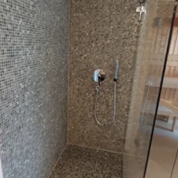 Kabina prysznicowa wyłożona mozaiką w odcieniach szarości i brązu, z widoczną słuchawką i deszczownicą, oddzielona szklaną ścianką od reszty łazienki z sauną w tle.