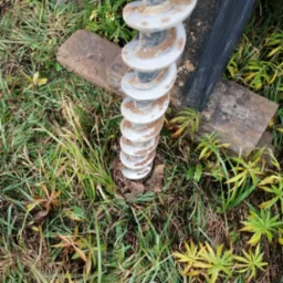 Wiertnica świdrowa zagłębiająca się w ziemię, widoczna w trawie z elementami drewnianymi podtrzymującymi konstrukcję.