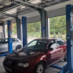 Dwa auta na podnośnikach w przestronnym warsztacie z otwartą bramą garażową. Jeden pojazd jest bordowy, drugi srebrny. Widoczne drabina i wyposażenie warsztatu.