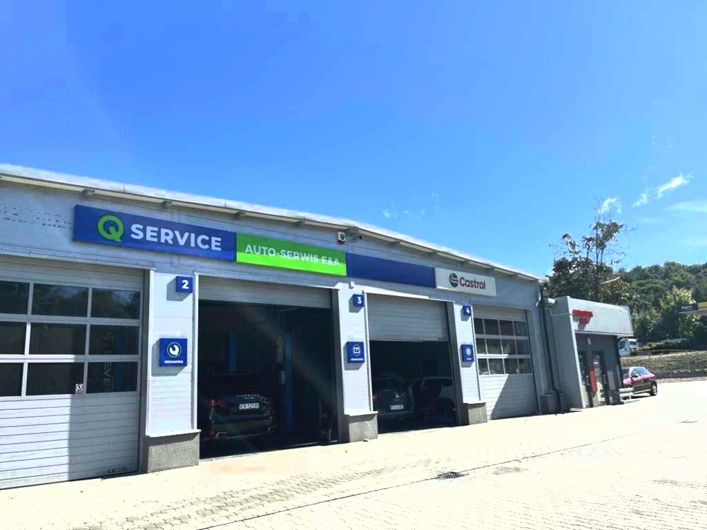 Zewnętrzny widok budynku auto serwisu z otwartymi bramami garażowymi, w których widoczne są samochody w trakcie naprawy. Szyldy Q Service i Castrol.