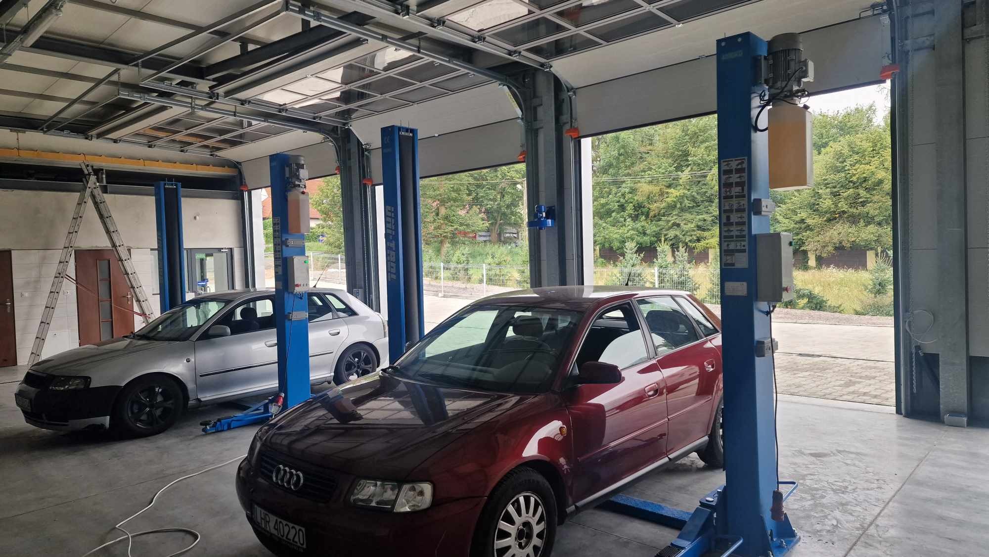 Dwa auta na podnośnikach w przestronnym warsztacie z otwartą bramą garażową. Jeden pojazd jest bordowy, drugi srebrny. Widoczne drabina i wyposażenie warsztatu.