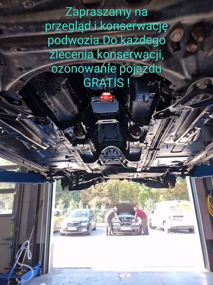 Podwozie auta po konserwacji na podnośniku, z tekstem o przeglądzie i ozonowaniu gratis. Mechanicy pracują przy innym aucie. Konserwacja podwozia.