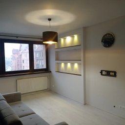 SERHII KOROLOV - Jasne wnętrze pokoju po remoncie: białe ściany, drewnopodobna podłoga, wnęki z oświetleniem, okno z widokiem na miasto. Na ścianie zegar i uchwyt do telewizora.