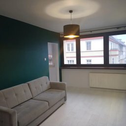 SERHII KOROLOV - Nowoczesne wnętrze: szmaragdowa ściana, beżowa sofa na jasnej podłodze, okno z widokiem na budynek, okrągła lampa sufitowa z ciepłym światłem.